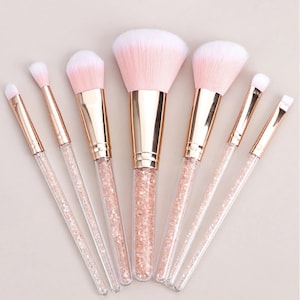 Könnte beinhalten: Ein Set aus sieben Make-up-Pinseln mit rosa und weißen Borsten. Die Pinsel haben transparente Griffe, die mit rosa Glitzer gefüllt sind, und goldfarbene Metallhülsen. Die Pinsel sind auf einer hellen Oberfläche angeordnet.