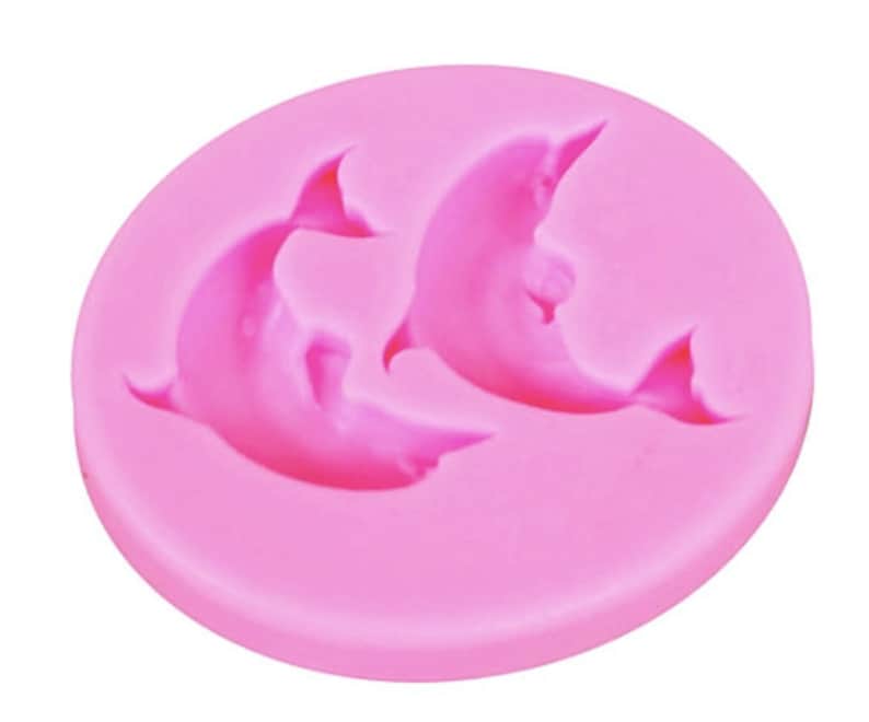 May include: Pink silicone mold with two dolphin shapes. The mold is designed for making cake decorations or other edible treats.