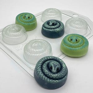 Könnte beinhalten: Eine transparente Kunststoffschale enthält mehrere seifenförmige Schlangen. Die Seifen sind in verschiedenen Farben erhältlich, darunter Grün, Blau und Weiß. Jede Seife hat ein detailliertes Schlangendesign, wobei Kopf und Körper gewickelt sind.