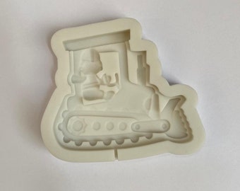 Puede incluir: Molde de silicona blanco de un bulldozer de dibujos animados con un conductor en la cabina. El molde está diseñado para hacer toppers de pastel u otras decoraciones comestibles.