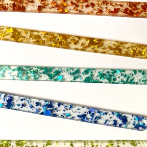 Peut inclure: Cinq b&acirc;tonnets acryliques transparents avec des paillettes de diff&eacute;rentes couleurs incrust&eacute;es &agrave; l'int&eacute;rieur. Les b&acirc;tonnets sont dispos&eacute;s verticalement de haut en bas avec les couleurs&nbsp;: orange, or, sarcelle, bleu et vert.