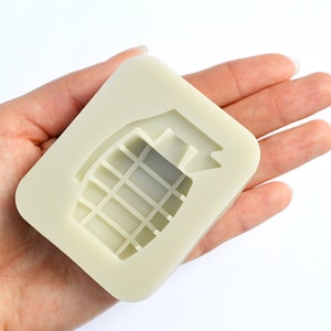 Puede incluir: Un molde de silicona beige claro con forma de granada de mano. El molde está diseñado para crear una réplica de una granada de mano.