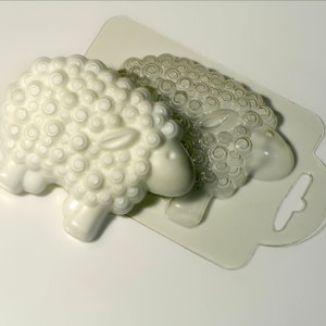 Puede incluir: Un molde de plástico blanco para hacer dos jabones con forma de oveja. El molde es transparente y muestra el detalle de la lana de la oveja.