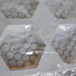 May include: A white silicone mould for crafting soaps or other items. The mould has a honeycomb design with a bee in each hexagonal cavity. The mould is packaged in clear plastic.