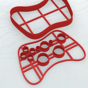 May include: Two red plastic cookie cutters in the shape of a video game controller. One cutter is a solid outline with a grid pattern, and the other has various circular and rectangular cutouts for buttons and controls. The cutters are on a white surface.