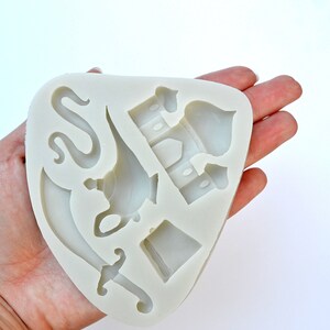 May include: A white silicone mold with five different shapes: a lamp, a sword, a snake, a hat, and a building with a dome. The mold is designed for making cake decorations.