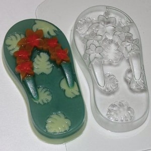 Könnte beinhalten: Eine transparente Plastikform zum Herstellen von Seife in Form einer Flip-Flop. Die Form hat ein tropisches Design mit grünen Blättern und orangefarbenen Blumen.