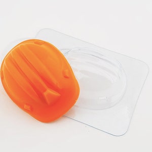 Op de afbeelding: Een oranje, helmvormige zeep ligt op een doorzichtige plastic mal. De zeep heeft een textuur met ribbels en een klein, driehoekig detail. De mal is transparant en de achtergrond is wit.
