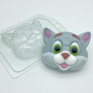 May include: A clear plastic mold of a cartoon cat face, with a gray, brown, and pink cat face made of soap in the mold.