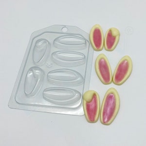 May include: A clear plastic mold with six cavities for making bunny ear-shaped treats. Three sets of bunny ears are shown, with white outer edges and pink inner details. Perfect for Easter or spring-themed baking.