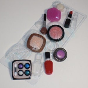 Peut inclure: Un moule en plastique transparent avec différentes formes pour réaliser des objets sur le thème du maquillage. Le moule comprend des formes pour le rouge à lèvres, les ombres à paupières, le fard à joues, le vernis à ongles et un pinceau à maquillage. Le moule est rempli d'objets de couleur rose, rouge et violette.