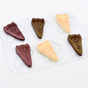 May include: A clear plastic mould containing six cat-shaped ice cream cones. The cones are made of chocolate in shades of brown and beige. Each cat has a smiling face.