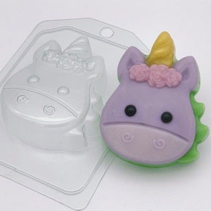 Puede incluir: Un molde de plástico transparente para hacer jabón con forma de unicornio. El molde se muestra con un jabón de unicornio morado con una crin verde y un cuerno dorado. El unicornio tiene flores rosas en la cabeza.