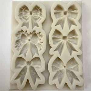May include: A light-colored silicone mold featuring various bow designs. The mold includes three bow shapes on the left and three on the right. The bows vary in style, with some having textured details and others appearing smooth.