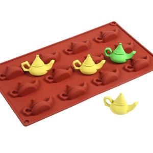 May include: A red silicone mold with 10 teapot-shaped cavities. There are three yellow and one green teapot-shaped candies in the mold.