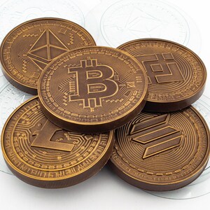 May include: A close-up of six chocolate coins featuring different cryptocurrency logos, including Bitcoin, Ethereum, Litecoin, and Binance Coin. The coins are arranged in a pile on a white surface.