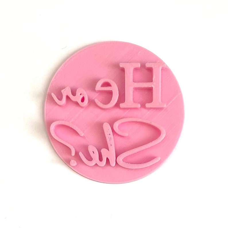 May include: Pink round rubber stamp with the text "He or She?" in white.