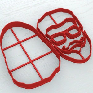 May include: Red plastic cookie cutters in the shape of a man's face with a mustache and glasses and a grid pattern.