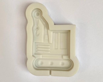Puede incluir: Molde de silicona blanco con forma de vehículo de construcción, una pequeña excavadora con un brazo levantado y un gancho. El molde está diseñado para hacer decoraciones comestibles.