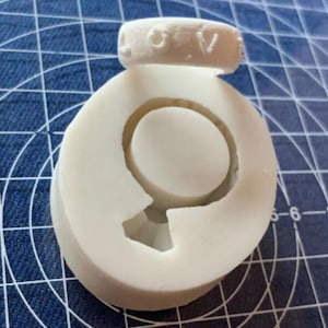 May include: White silicone mold for making a ring with a raised ring shape in the center. The mold is sitting on a blue grid surface.