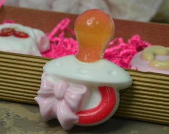 May include: A white baby dummy soap with a pink bow and a red handle. The dummy is sitting on a brown and pink surface.
