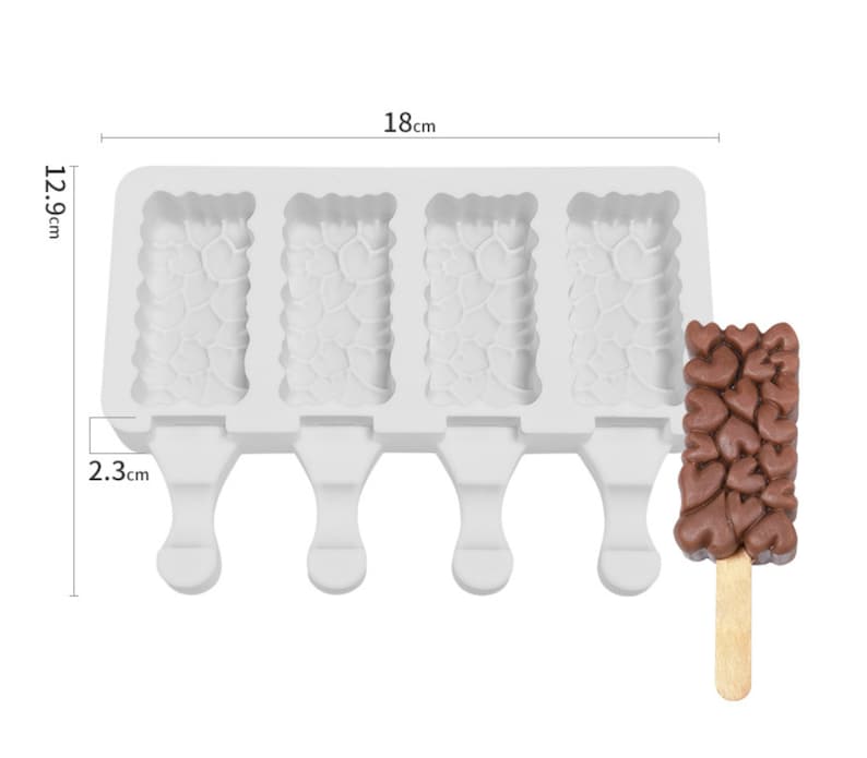 May include: White silicone mold with four rectangular cavities for making popsicles. Each cavity has a heart-shaped design. The mold has four handles for holding the popsicles. A chocolate popsicle with a wooden stick is shown next to the mold. The popsicle is also heart-shaped.
