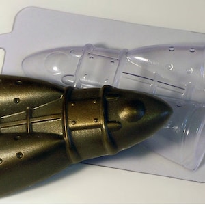 May include: A bronze-colored rocket-shaped mold for crafting. The mold is made of a metallic-looking material and is packaged in a clear plastic container. The mold has detailed features, including rivets and panel lines, suggesting a vintage or industrial design.