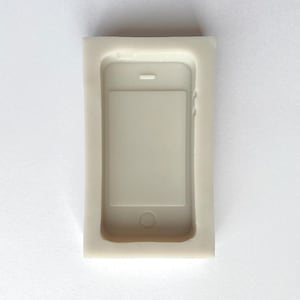 May include: A white silicone mold shaped like a smartphone. The mold has a rectangular shape with rounded corners and a raised outline of a smartphone screen, camera, and home button.