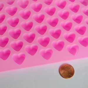 Puede incluir: Un molde de silicona rosa con m&uacute;ltiples cavidades en forma de coraz&oacute;n. Se coloca una moneda junto al molde para comparar el tama&ntilde;o. El molde est&aacute; dise&ntilde;ado para crear peque&ntilde;os objetos con forma de coraz&oacute;n, probablemente para hornear o manualidades.