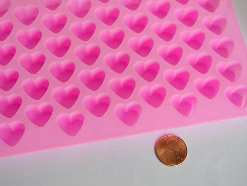 Puede incluir: Un molde de silicona rosa con m&uacute;ltiples cavidades en forma de coraz&oacute;n. Se coloca una moneda junto al molde para comparar el tama&ntilde;o. El molde est&aacute; dise&ntilde;ado para crear peque&ntilde;os objetos con forma de coraz&oacute;n, probablemente para hornear o manualidades.