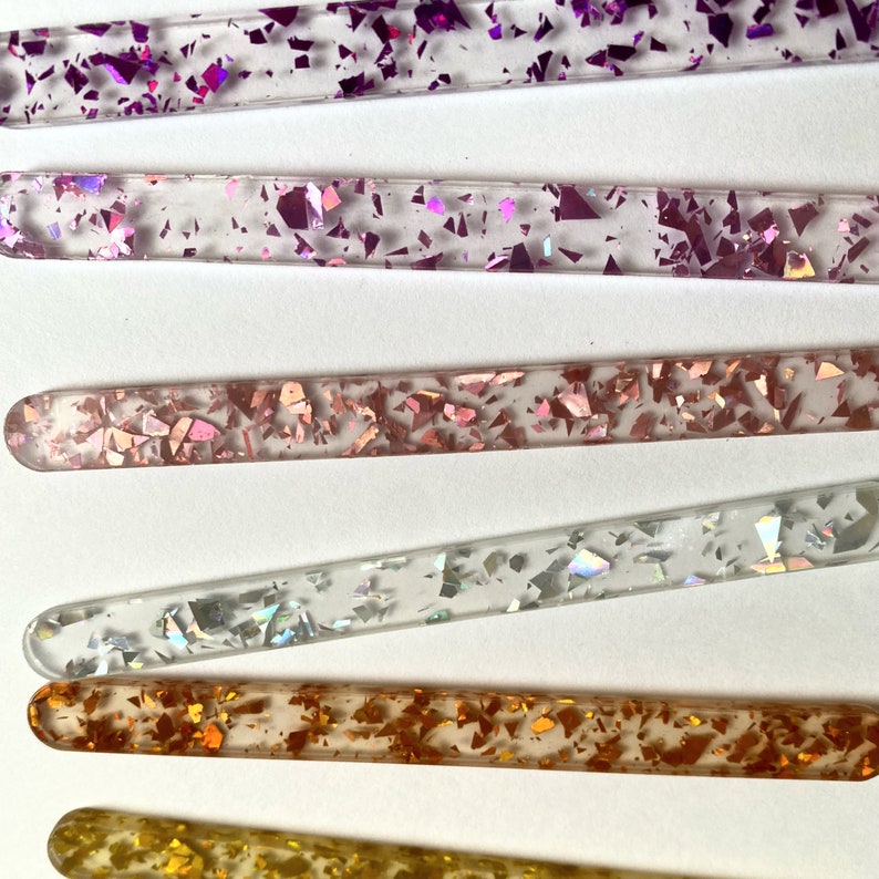 Peut inclure: Cinq b&acirc;tonnets en verre transparent avec des paillettes de diff&eacute;rentes couleurs incrust&eacute;es &agrave; l'int&eacute;rieur. Les b&acirc;tonnets sont dispos&eacute;s verticalement, le b&acirc;tonnet &agrave; paillettes violettes en haut et le b&acirc;tonnet &agrave; paillettes dor&eacute;es en bas.