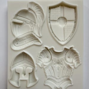 May include: A white silicone mold featuring four medieval-themed designs: a knight's helmet, a shield, a breastplate, and a second helmet. The mold is designed for creating decorative elements for cakes, crafts, or other projects.