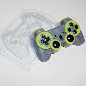 May include: A grey and green plastic mould for making a video game controller shaped soap. The mould is made of clear plastic and has a detailed design of a video game controller.