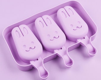 May include: A lilac-coloured popsicle mould featuring three bunny-shaped cavities. Each bunny has a smiling face and long ears. The mould includes a tray and handles for easy removal of the frozen treats.
