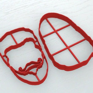 May include: Two red plastic cookie cutters, one shaped like a face with a mustache and the other shaped like a rectangle with a grid pattern.