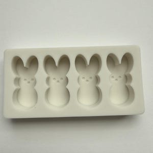 May include: A white silicone mold with four bunny-shaped cavities. Each bunny has a rounded body, long ears, and three small dots for facial features. The mold is rectangular and sits on a white surface.