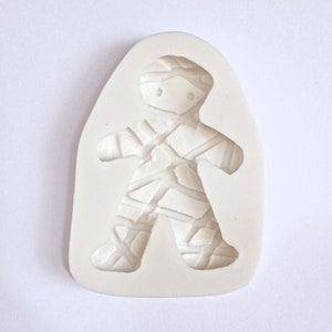 May include: White silicone mold shaped like a mummy. The mummy is standing with its arms outstretched and has a simple face with two eyes.