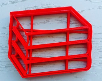 May include: A red plastic cookie cutter shaped like a stack of three rectangular boxes. The cutter has a detailed design with lines and edges.