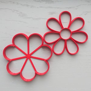 Könnte beinhalten: Zwei rote Plastik-Ausstecher in Form von Blumen. Ein Ausstecher ist eine einfache 8-blättrige Blume, der andere eine komplexere 10-blättrige Blume.
