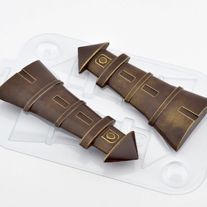 May include: Two brown and gold lighthouse-shaped chocolate molds. The molds are made of clear plastic and are sitting on a white surface.