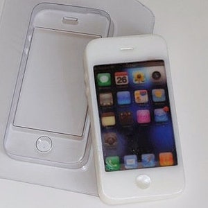May include: White miniature smartphone with a screen displaying colorful app icons. The phone has a home button and a speaker at the top. A clear plastic mold of the phone is next to it.