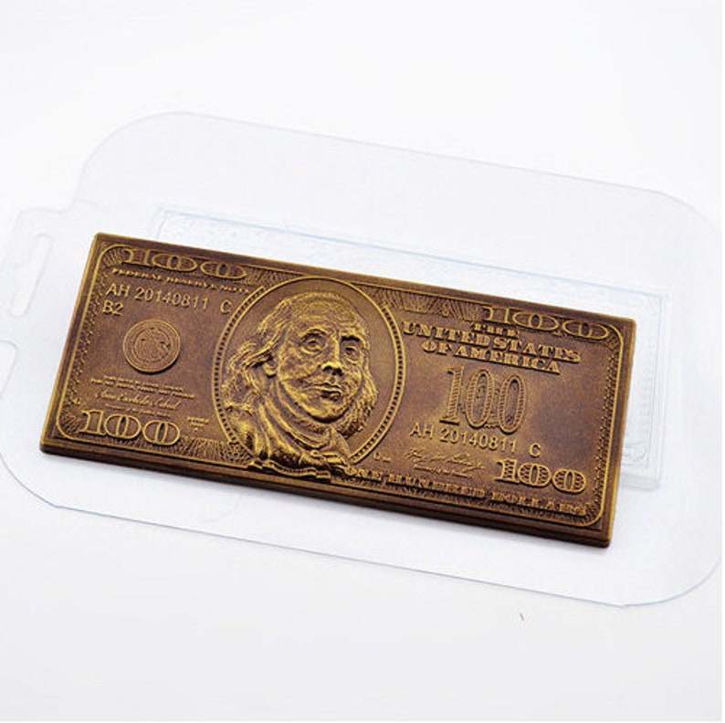 May include: A bronze-colored rectangular object resembling a U.S. $100 bill. The design includes a portrait, the text "The United States of America," and the number "100." The bill is encased in a clear plastic sleeve.