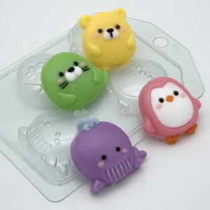 Op de afbeelding: Een transparante plastic mal met vier holtes voor het maken van kleine dierensaponnen. De mal is gevuld met vier afgewerkte zepen: een gele beer, een groene zeehond, een roze pinguïn en een paarse walvis.