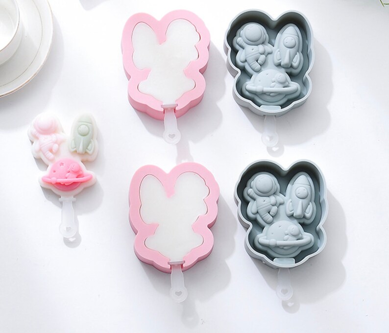 Puede incluir: Dos juegos de moldes de paletas de hielo de silicona. Un juego es rosa y presenta un dise&ntilde;o de conejo. El otro juego es gris y presenta un dise&ntilde;o de astronauta, cohete y planeta.