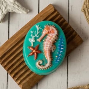 May include: A teal blue soap bar with a seahorse, starfish, and coral design. The soap is on a wooden soap dish.