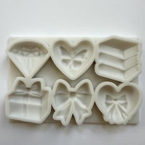 Może przedstawiać: Biała silikonowa forma z sześcioma różnymi kształtami: diamentem, sercem z motylem, stos książek, pudełkiem prezentowym z kokardą, dużą kokardą i sercem z kokardą. Kształty są wypukłe i szczegółowe.