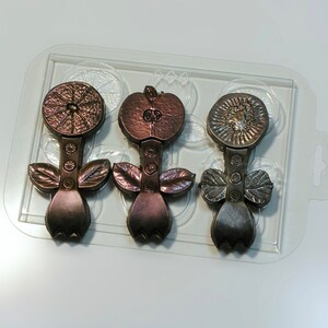 May include: Three chocolate molds in a clear plastic tray. The molds are shaped like flowers with a fruit design on top. The fruit designs are a lemon, an apple, and a sunflower. The chocolate is a dark brown color.