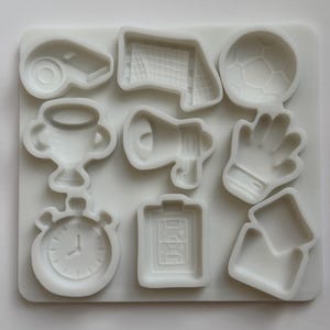 May include: A white silicone mold featuring nine distinct sports-themed cavities, including a whistle, soccer goal, soccer ball, trophy, megaphone, goalkeeper glove, stopwatch, soccer field clipboard, and a set of penalty cards.