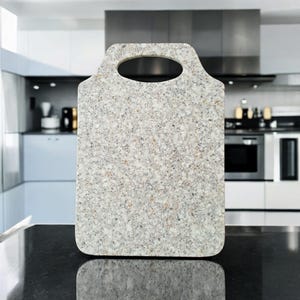 Puede incluir: Una tabla de cortar rectangular con asa. La tabla es de color gris claro con motas de gris más oscuro y marrón. El asa tiene una abertura ovalada. La tabla está sobre una encimera negra en una cocina.