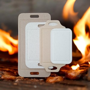 May include: Four rectangular cutting boards in varying sizes and shades of beige and white are stacked together. The boards have a speckled texture and a handle or cutout for hanging. The background features a blurred image of a fire.